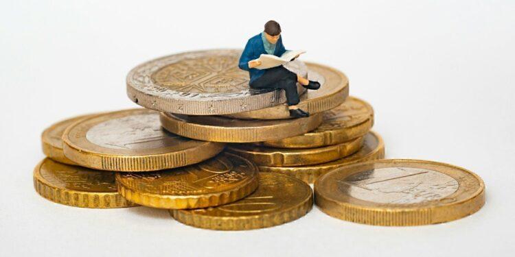 Miniature person sitting on stack of coins reading newspaper