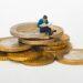 Miniature person sitting on stack of coins reading newspaper