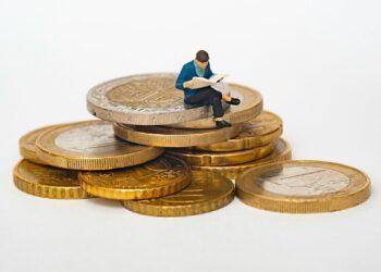 Miniature person sitting on stack of coins reading newspaper