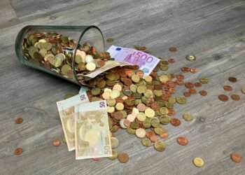 A glass jar with euro coins and banknotes spilled on a wooden surface, representing finance and savings.