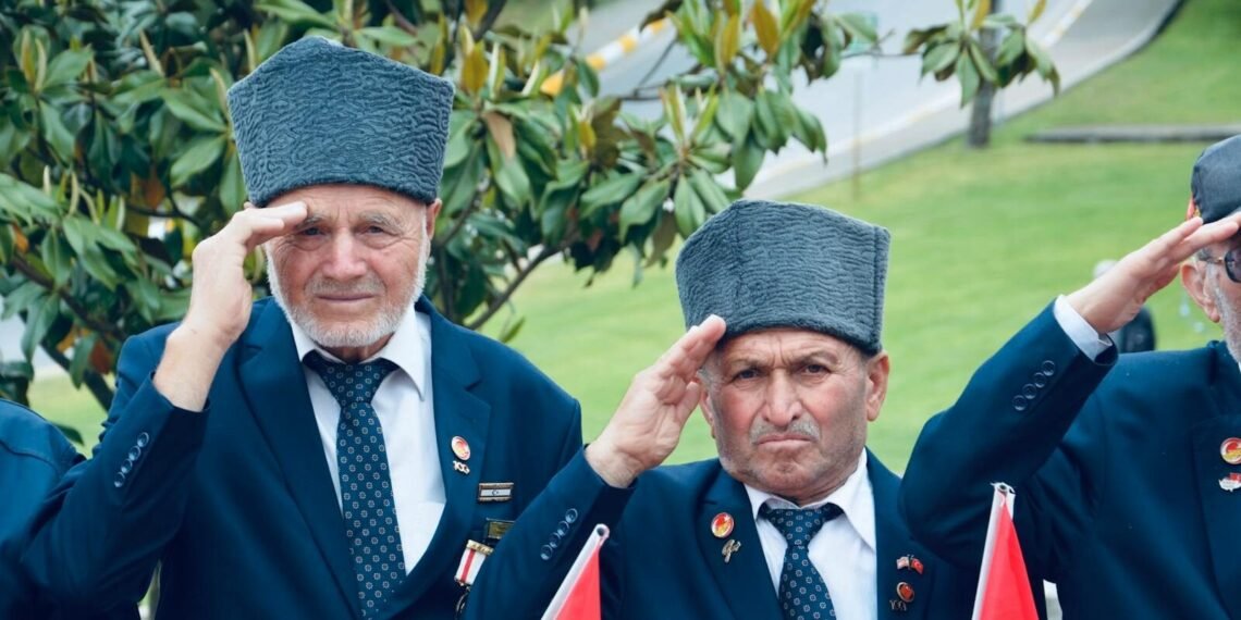 Two elderly veterans saluting during an outdoor ceremony with flags in Balıkesir, Türkiye.
