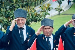 Two elderly veterans saluting during an outdoor ceremony with flags in Balıkesir, Türkiye.